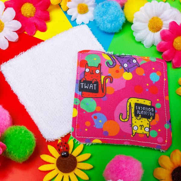 a bright pink Reusable Makeup Remover Pad with coloured circles on it and smiley cats holding black signs with sweary words on them illustrated by Kate Abey. The pad is sat on a green backdrop with colourful flowers scattered round