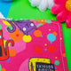 a bright pink Reusable Makeup Remover Pad with coloured circles on it and smiley cats holding black signs with sweary words on them illustrated by Kate Abey. Next to it is another makeup remover pad sat face down to show the plain white side of the pad. The pad is sat on a green backdrop with colourful flowers scattered round