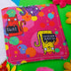 a bright pink Reusable Makeup Remover Pad with coloured circles on it and smiley cats holding black signs with sweary words on them illustrated by Kate Abey. Next to it is another makeup remover pad sat face down to show the plain white side of the pad. The pad is sat on a green backdrop with colourful flowers scattered round