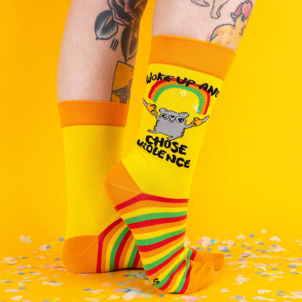 A side view of yellow socks with a bold design of a hamster with chainsaws and the words “Woke Up and Chose Violence,” paired with striped soles, modelled on tattooed legs against a vibrant yellow backdrop.