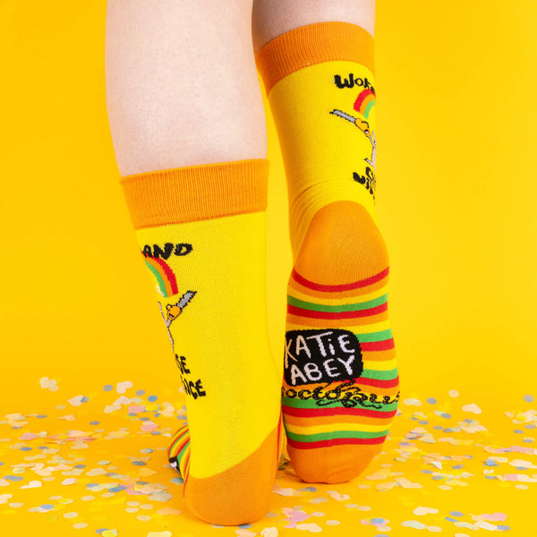 A back view of bright yellow socks with orange cuffs and toes, featuring a quirky hamster wielding chainsaws under a rainbow, and “Katie Abey” branding on the soles, set against a confetti-filled yellow background.