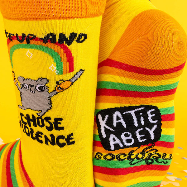 A close-up of the sock design showing the hamster, rainbow, and cheeky slogan “Woke Up and Chose Violence,” alongside colourful striped soles, with confetti scattered in the background.