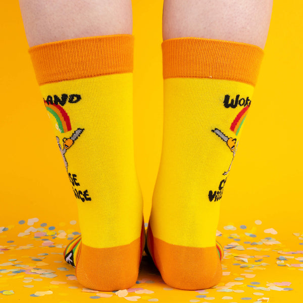 A rear view of the socks showing the hamster design and slogan “Woke Up and Chose Violence” on both legs, with confetti scattered on the floor against a yellow background.