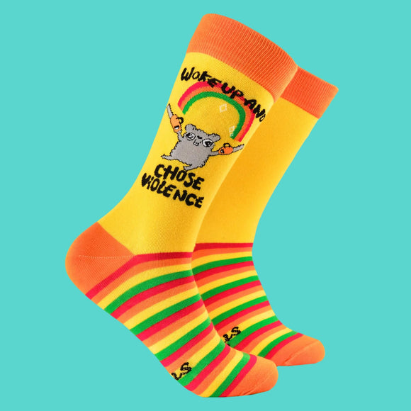 Bright yellow socks by Katie Abey with the slogan "Woke Up and Chose Violence" featuring a quirky grey hamster holding chainsaws under a sparkly rainbow. The socks have rainbow-striped soles and orange accents on the toes, heels, and cuffs. Perfect for bold, fun fashion.