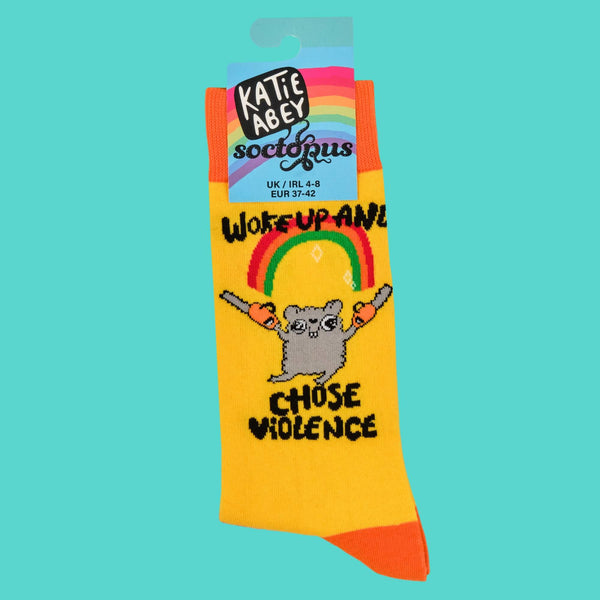 Katie Abey yellow socks with vibrant orange toes and cuffs, featuring a grey hamster holding chainsaws under a sparkly rainbow with "Woke Up and Chose Violence" text. Displayed with the Socktopus-branded label, highlighting Katie's fun, weird and playful small business aesthetic.