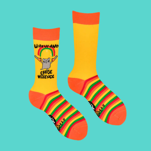 A flat-lay of Katie Abey's bright yellow socks with orange accents and rainbow-striped soles. Features a grey hamster with chainsaws under a sparkly rainbow, with "Woke Up and Chose Violence" text. Perfect for lovers of colourful, quirky accessories with a touch of chaos.