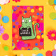 Fun A6 greeting card postcard with green cat holding funny sweary sign and smiley face, on colourful background designed by Katie Abey in the UK. It is on a yellow background with confetti, fake daisies and pom poms.