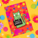 Fun A6 greeting card postcard with green cat holding funny sweary sign and smiley face, on colourful background designed by Katie Abey in the UK. It is on a yellow background with confetti, fake daisies and pom poms.