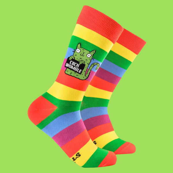 Bright rainbow-striped socks by Katie Abey featuring a quirky green cat character holding a "Cock Womble" sign. Perfect for those who love bold, weird designs with a humorous twist. Fun gift idea from a small business encouraging individuality and creativity.