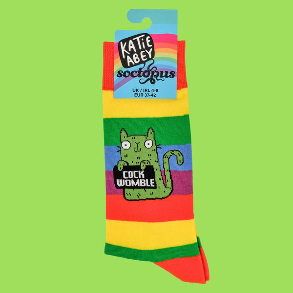 Rainbow socks by Katie Abey, showcasing packaging with a green cat holding a "Cock Womble" sign. A playful, bold accessory for fans of weird and unique styles. Designed by a small UK illustrator promoting creativity and embracing your weirdness.