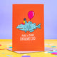 The birthday Cod a6 greeting card designed and printed in the UK by Katie Abey. The card is stood upright on the brown envelope it comes with, blue scale paper and pink sweets surrounding it on a white background. The front cover is an orange background with a blue cod fish wearing a rainbow birthday hat and holding a red balloon pulling a grimaced face. The text underneath the fish reads 'Here is your birthday cod'.
