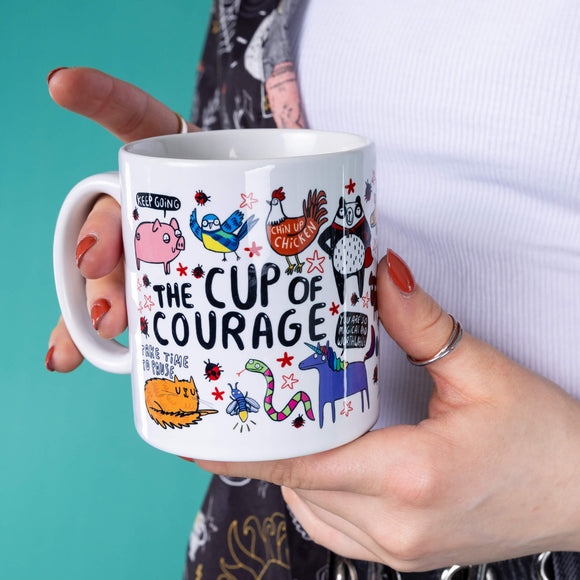 Amy is holding the cup of courage by Katie Abey in her hands in a studio. There are lots of Katies characters on the mug including unicorns, cats, snakes, pigs, chickens and more.