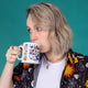 Amy is sipping tea out of the cup of courage by Katie Abey in her hands in a studio. There are lots of Katies characters on the mug including unicorns, cats, snakes, pigs, chickens and more.