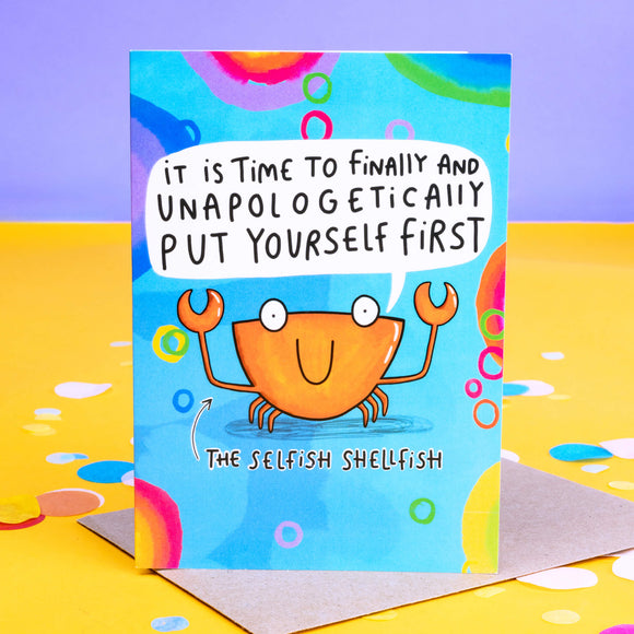Blue coloured greeting card with rainbow designs and crab illustration on the front with white speech bubble above crabs mouth saying 'it is time to finally and unapologetically put yourself first'. There is also black writing under the crab saying 'The Selfish Shellfish' with an arrow up to the crab.
