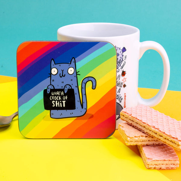 A coaster with a purple smiling cat illustration by Katie Abey holding a sign saying 'what a crock of shit' on a rainbow background.
