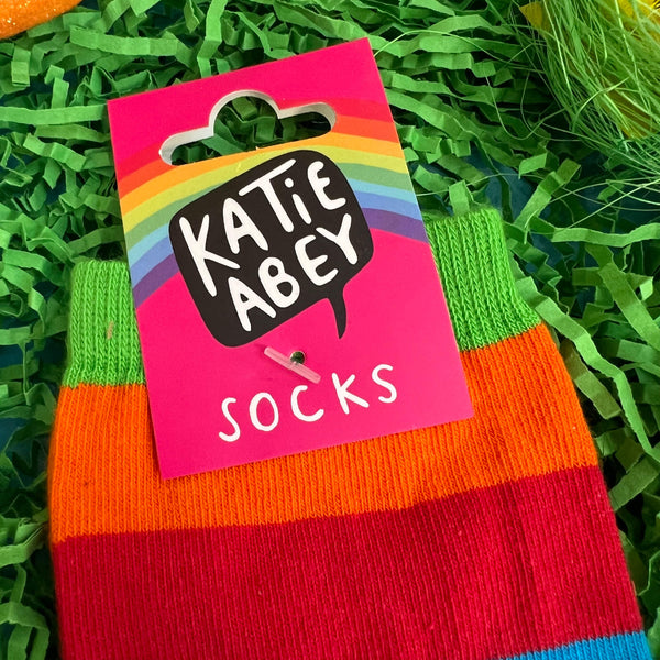 pink cardboard label with rainbow and Katie Abey logo on attached to the What A Crock Of Sh*t Rainbow Sweary Cat Socks laying on some grass in the sun