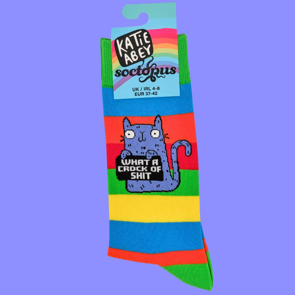 Close-up of rainbow-striped socks featuring Katie Abey's whimsical blue cat holding a sign reading, "What a crock of shit." Packaged with Katie Abey branding, this vibrant design celebrates the weird and wonderful style of this independent illustrator.