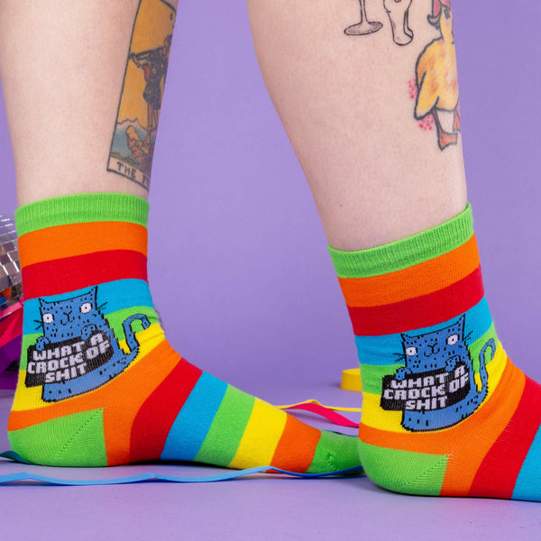 A model wearing Katie Abey socks with a blue cat holding a sign saying what a crock of shit. They are striped in rainbow colours and lovely and vibrant. The model is stood on a purple floor with purple with disco balls and ribbons