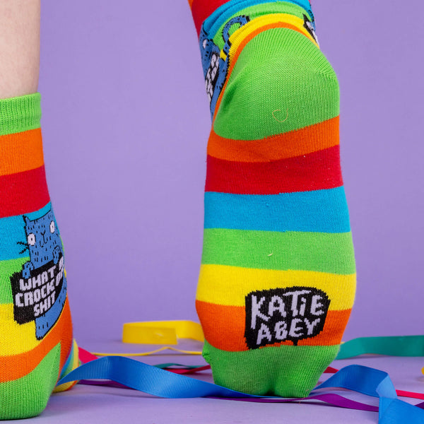 A model wearing Katie Abey socks with Katie Abey logo on the bottom. They are striped in rainbow colours and lovely and vibrant. The model is stood on a purple floor with purple with disco balls and ribbons