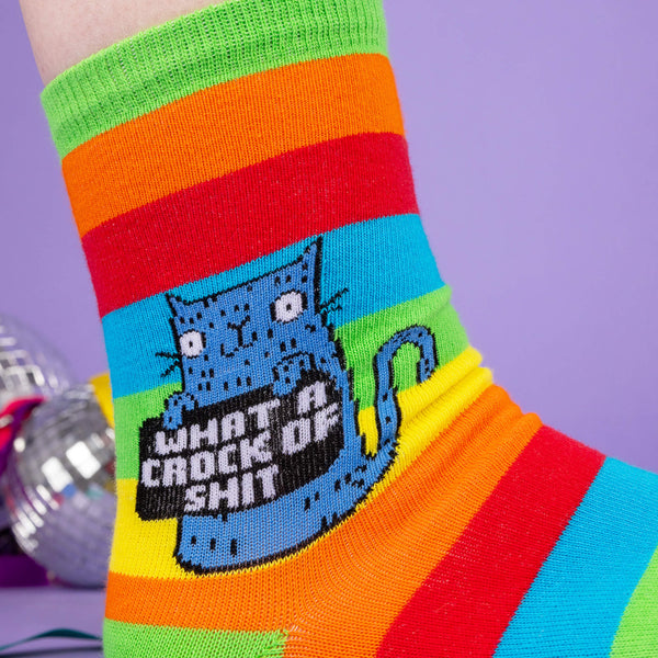 close up of model wearing Katie Abey socks with a blue cat holding a sign saying what a crock of shit. They are striped in rainbow colours and lovely and vibrant. The model is stood on a purple floor with purple with disco balls and ribbons