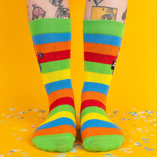 A pair of feet wearing bright rainbow-striped socks featuring a sassy purple cat holding a sign reading “What a crock of sh*t.” The socks have a vibrant green cuff and are surrounded by colourful confetti on a yellow background.