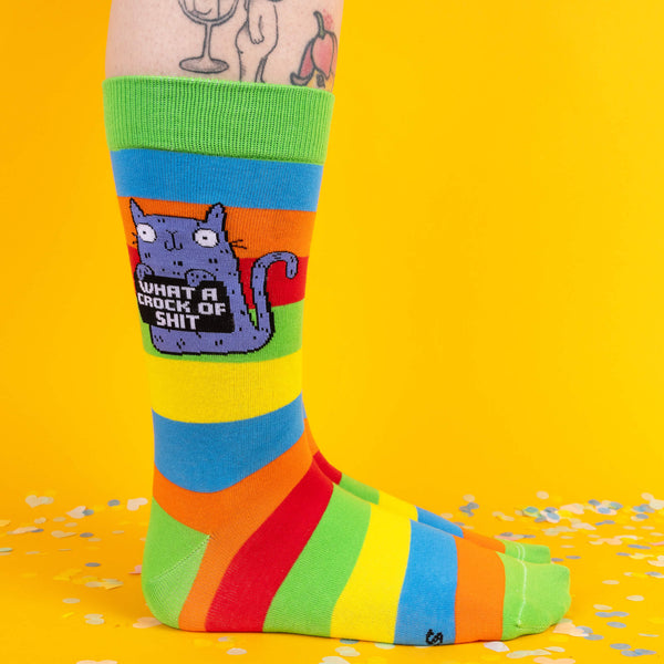 Side view of rainbow-striped socks, highlighting the purple cat design with the text “What a crock of sh*t.” The green cuff and bold stripes add a playful touch against the bright yellow backdrop.