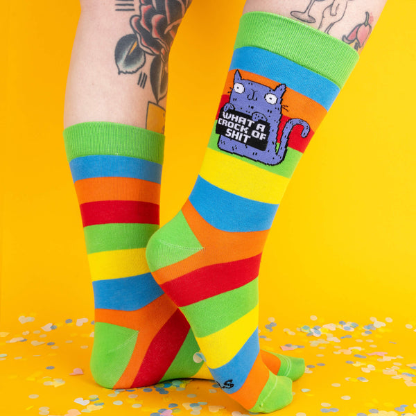  Side view of rainbow-striped socks, highlighting the purple cat design with the text “What a crock of sh*t.” The green cuff and bold stripes add a playful touch against the bright yellow backdrop.