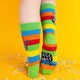 Back view of the rainbow-striped socks, showing the vibrant design with a green cuff, and a small section of the purple cat’s tail. Confetti is scattered on the yellow surface below.