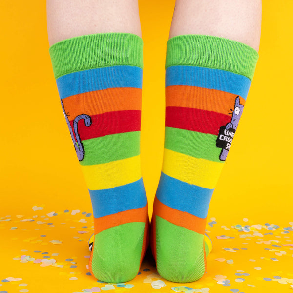 Back view of the rainbow-striped socks, showing the vibrant design with a green cuff, and a small section of the purple cat’s tail. Confetti is scattered on the yellow surface below.