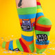 A playful pose showing the side and bottom of the socks. The soles feature the “Katie Abey x Soctopus” logo, with the purple cat design standing out boldly against the colourful stripes.