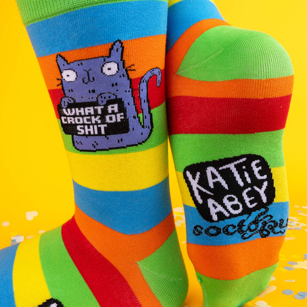 A playful pose showing the side and bottom of the socks. The soles feature the “Katie Abey x Soctopus” logo, with the purple cat design standing out boldly against the colourful stripes.