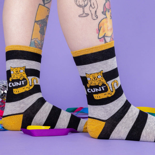 A white model with tattooed legs is wearing a pair of black and grey striped socks with gold toes, heel and band around the top. There is a gold cat illustration on them by Katie Abey holding a sign saying cunt