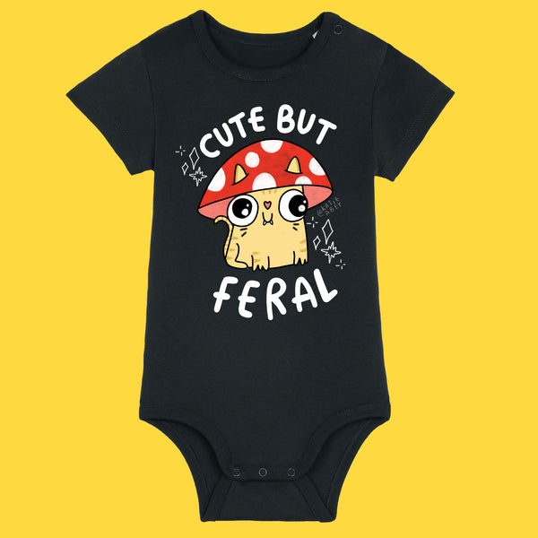 Black baby grow featuring the Cute But Feral Cat illustration by Katie Abey. The design shows a wide eyed cartoon cat under a red spotted mushroom with bold white Cute But Feral text. Made from soft organic cotton with popper fastenings for easy changes.