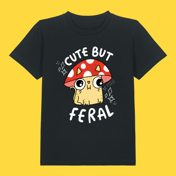 Front view of the Cute But Feral Cat Kids T Shirt by Katie Abey in black. Kids organic cotton tee featuring a cute feral cat illustration under a red mushroom with the slogan Cute But Feral, photographed on a bright yellow background.