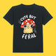 Front view of the Cute But Feral Cat Kids T Shirt by Katie Abey in black. Kids organic cotton tee featuring a cute feral cat illustration under a red mushroom with the slogan Cute But Feral, photographed on a bright yellow background.