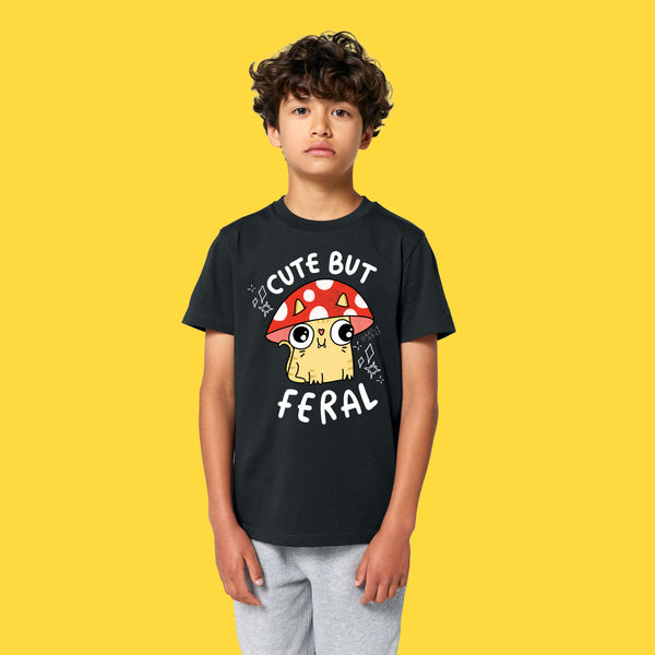 Front view of a child wearing the Cute But Feral Cat Kids T Shirt by Katie Abey in black. Features Katie Abey’s Cute But Feral cat artwork with mushroom illustration and bold white lettering on a soft organic cotton kids T shirt.