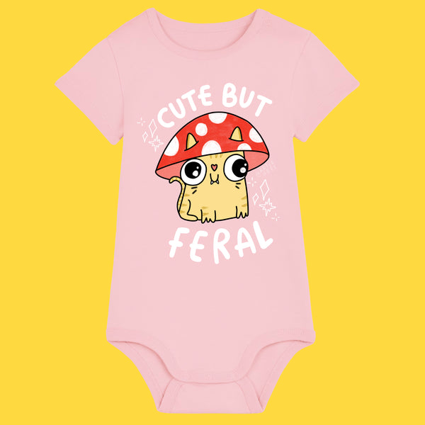 Pink baby grow featuring the Cute But Feral Cat illustration by Katie Abey. The playful design shows a wide eyed cartoon cat under a red spotted mushroom with bold Cute But Feral text. Made from organic cotton with shoulder snaps and bottom poppers for easy changing.
