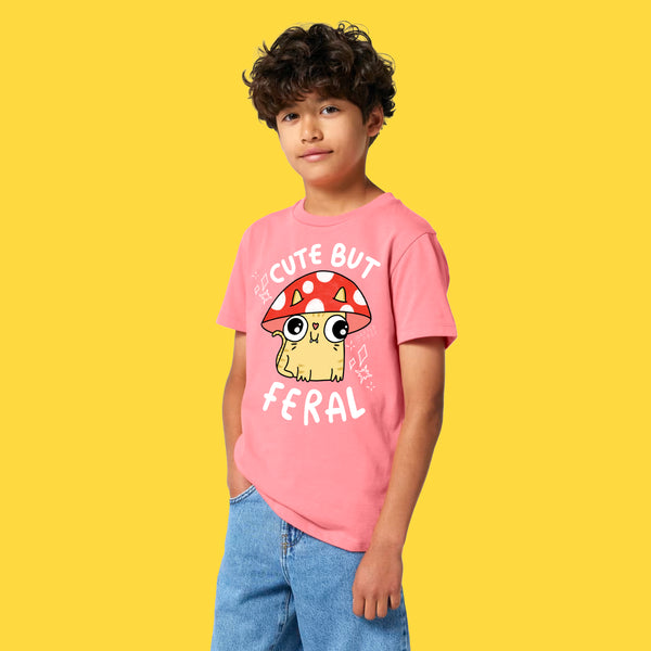 Front view of a child wearing the Cute But Feral Cat Kids T Shirt by Katie Abey in pink joy. Features Katie Abey’s playful Cute But Feral cat artwork with mushroom illustration and bold white text on an organic cotton kids tee.