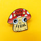 Close-up of the Cute But Feral Mushroom Cat enamel pin by Katie Abey, showing a yellow cartoon cat with a red polka dot toadstool hat and big wide eyes. The mushroom cap reads “Cute But Feral” in bold black text.