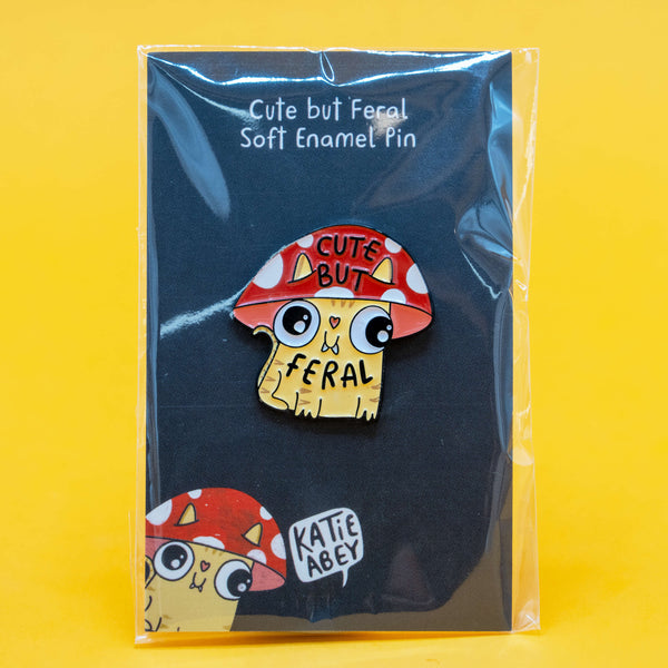 Photo of Katie Abey’s Cute But Feral Mushroom Cat enamel pin in packaging, displayed against a black backing card with a red toadstool cat design. The quirky illustrated pin features a yellow cat with a red mushroom cap that says “Cute But Feral”.