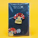 Photo of Katie Abey’s Cute But Feral Mushroom Cat enamel pin in packaging, displayed against a black backing card with a red toadstool cat design. The quirky illustrated pin features a yellow cat with a red mushroom cap that says “Cute But Feral”.