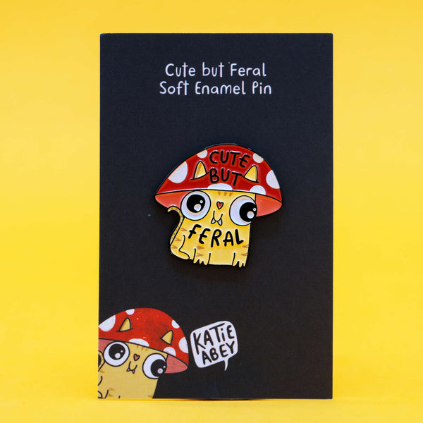 Katie Abey’s Cute But Feral Mushroom Cat enamel pin on a display card with branding. The illustrated yellow cat wears a red mushroom cap with ears and “Cute But Feral” text, photographed on a bright yellow background.