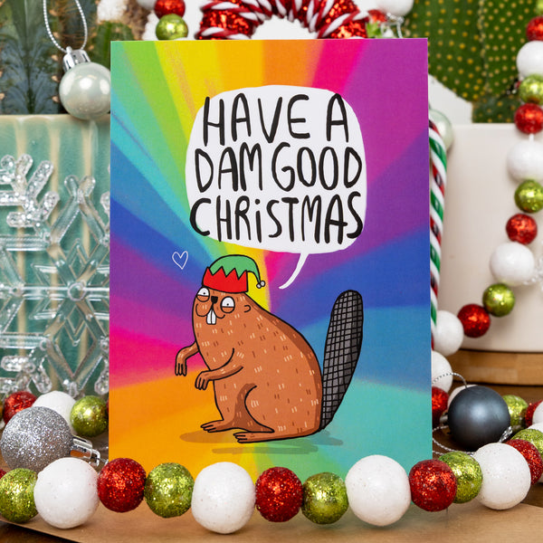 A fun, colourful Christmas card showing a cartoon beaver wearing a green and red elf hat. The speech bubble reads, "Have a dam good Christmas." Background features a rainbow gradient with festive decor around the card.