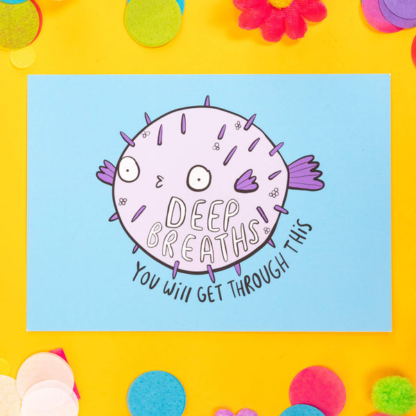 Postcard with a puffer fish looking like a spooky balloon with 'deep breaths you will get through this' on a blue background. the postcard is laid on a yellow background with colourful confetti, daisies and pom poms