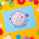 Postcard with a puffer fish looking like a spooky balloon with 'deep breaths you will get through this' on a blue background. the postcard is laid on a yellow background with colourful confetti, daisies and pom poms