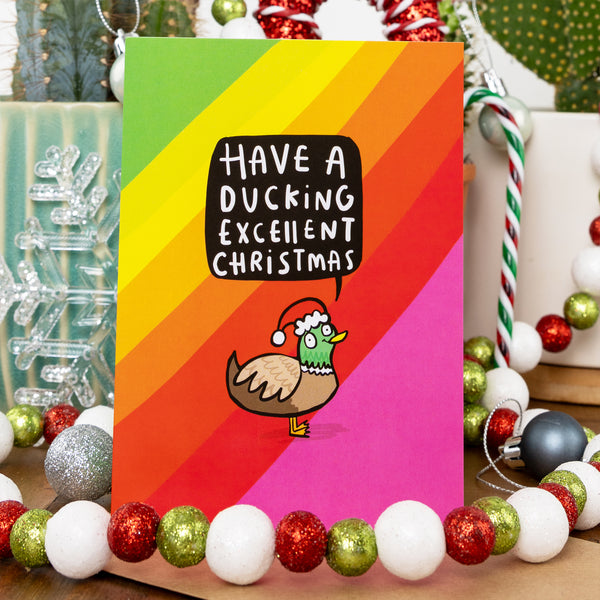 A vibrant Christmas card with a bold rainbow gradient background featuring a cartoon duck wearing a Santa hat. The card humorously reads, 'Have a ducking excellent Christmas' in a playful speech bubble. The scene is surrounded by festive decorations, including red, green, and white glittery baubles, a silver snowflake, and a striped candy cane