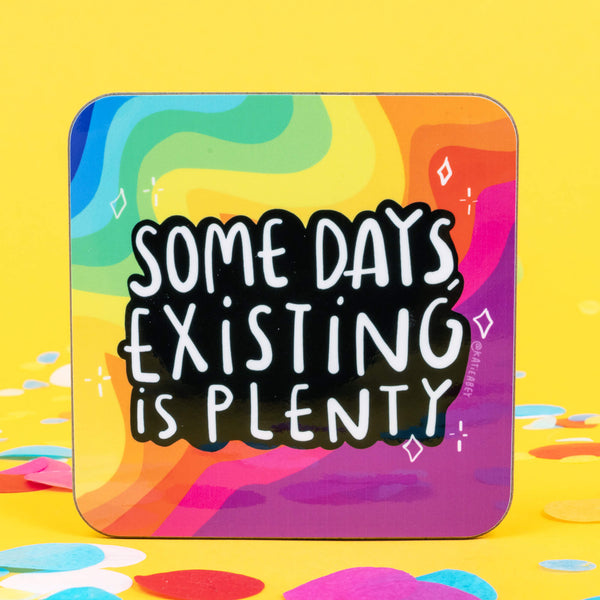 A coaster designed and illustrated by Katie Abey with rainbow background and text saying, 'some days, existing is plenty' 