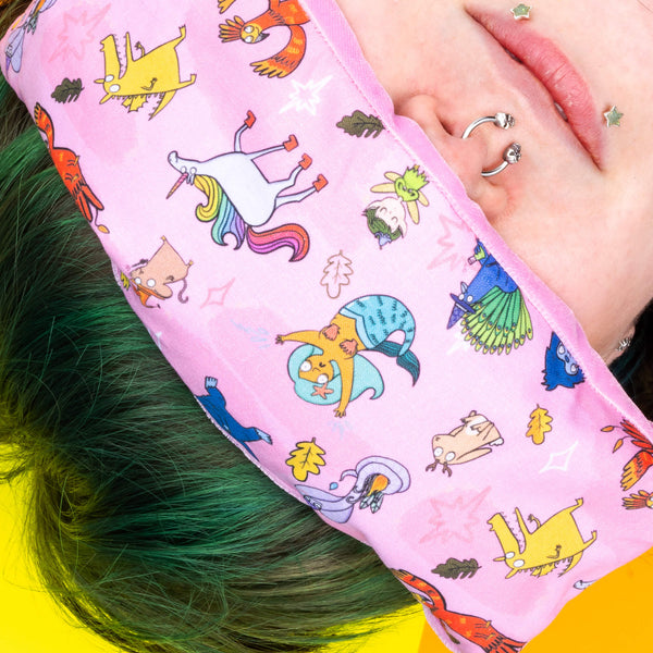 Faeryn a green haired human with facial piercings has a pink eye mask on with Katie Abey Happiness Enchanters characters on including unicorns, mermaids and dragons