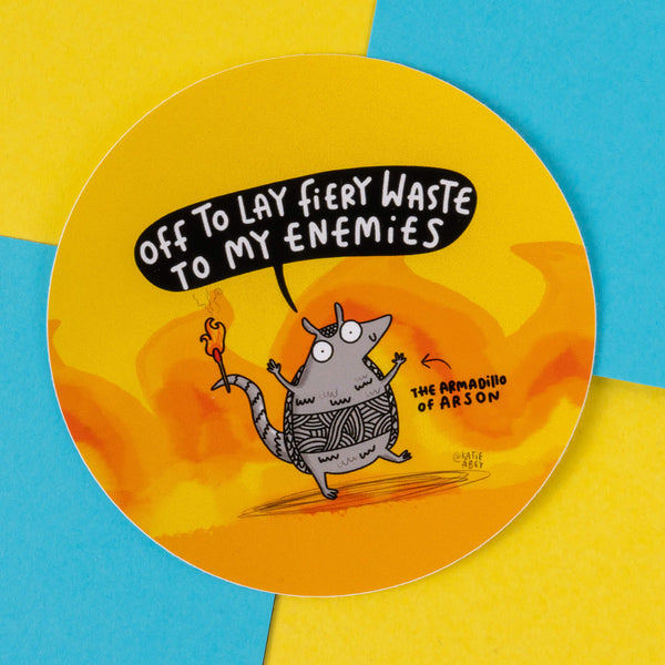 A circular sticker featuring an illustrated armadillo holding a small flame. The character says, "Off to lay fiery waste to my enemies" with the caption "The Armadillo of Arson" next to it. The background is vibrant with yellow and orange flames, and blue accents. This artwork is by Katie Abey, an illustrator known for her quirky and humorous designs that encourage embracing your weirdness.