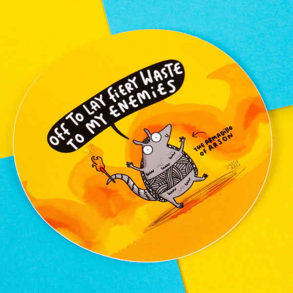 A circular sticker displaying an armadillo character holding a flame, saying, "Off to lay fiery waste to my enemies." The text "The Armadillo of Arson" appears beside it. The vibrant background features yellow and orange flames with blue accents. Designed by Katie Abey, a small business and illustrator known for promoting self-expression and weirdness through playful art.